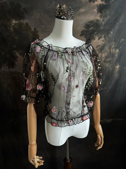 Mannequin wearing a gothic black sheer mesh puff sleeve blouse with embroidered botanicals and a ruffle hem, on a forest-themed art history background