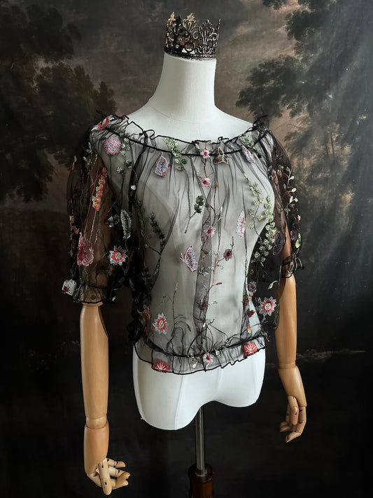 Mannequin wearing a gothic black sheer mesh puff sleeve blouse with embroidered botanicals and a ruffle hem, on a forest-themed art history background