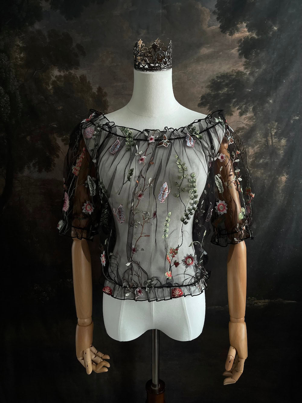 Mannequin wearing a gothic black sheer mesh puff sleeve blouse with embroidered botanicals and a ruffle hem, on a forest-themed art history background