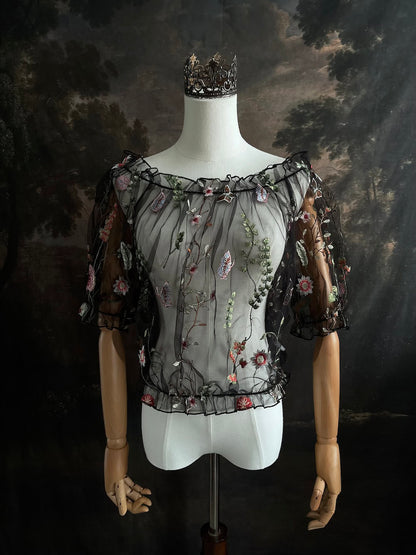 Mannequin wearing a gothic black sheer mesh puff sleeve blouse with embroidered botanicals and a ruffle hem, on a forest-themed art history background