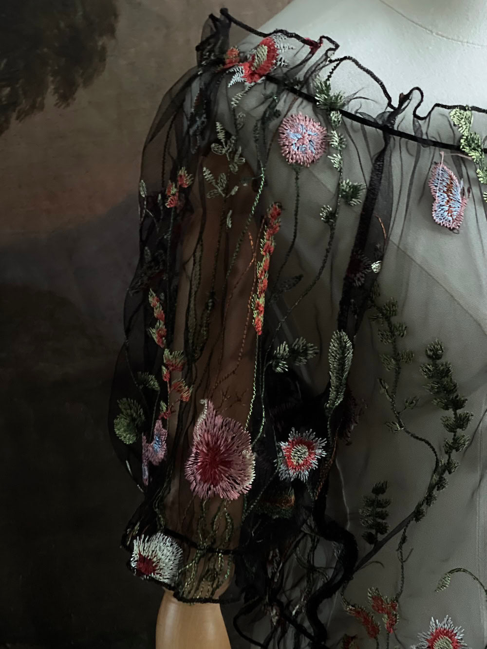 details of a gothic black sheer mesh puff sleeve blouse with embroidered botanicals and a ruffle hem, on a forest-themed art history background