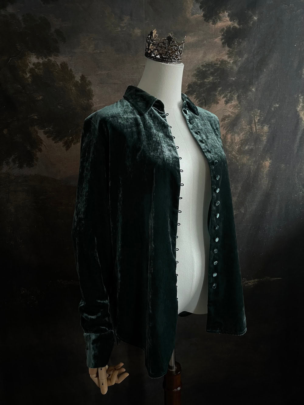 Mannequin wearing a dark green velvet button up vintage medieval revival style collared shirt against a dark, forest-themed art history background