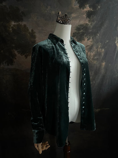 Mannequin wearing a dark green velvet button up vintage medieval revival style collared shirt against a dark, forest-themed art history background