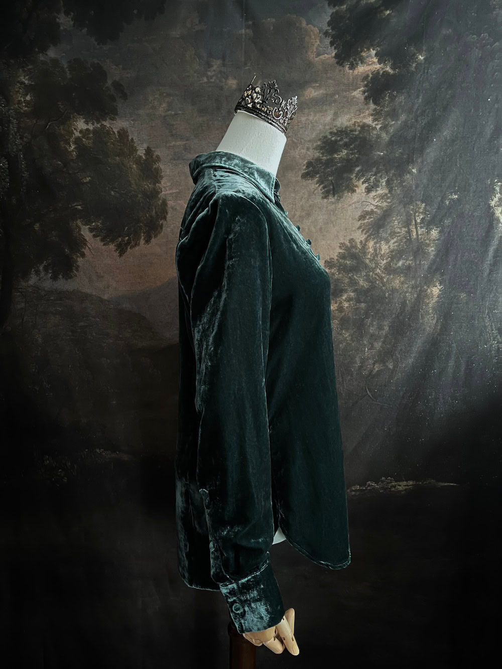 Mannequin wearing a dark green velvet button up vintage medieval revival style collared shirt against a dark, forest-themed art history background
