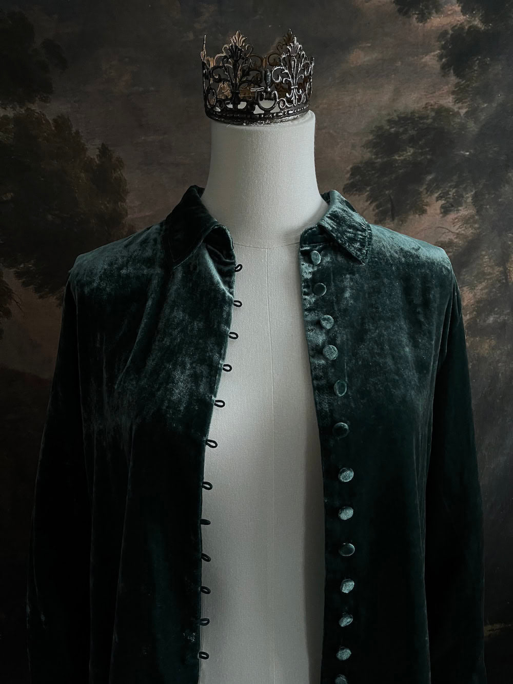 Mannequin wearing a dark green velvet button up vintage medieval revival style collared shirt against a dark, forest-themed art history background