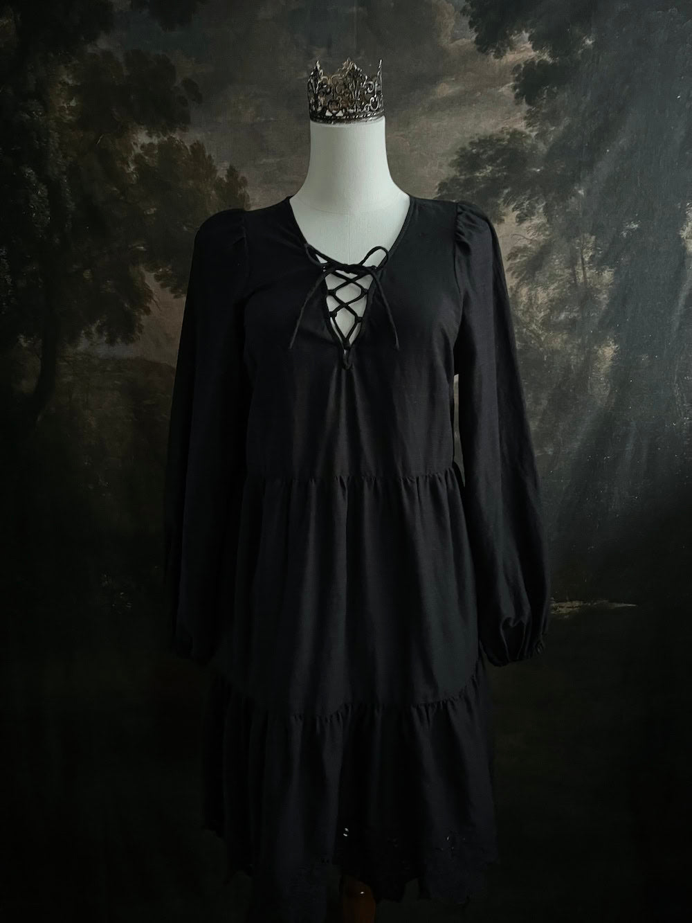 Historically inspired witchy gothic black tiered mini dress with corset lace up bodice and long bishop sleeves on a mannequin in front of a dark art history backdrop