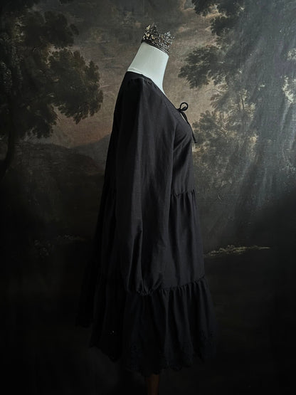 Historically inspired witchy gothic black tiered mini dress with corset lace up bodice and long bishop sleeves on a mannequin in front of a dark art history backdrop
