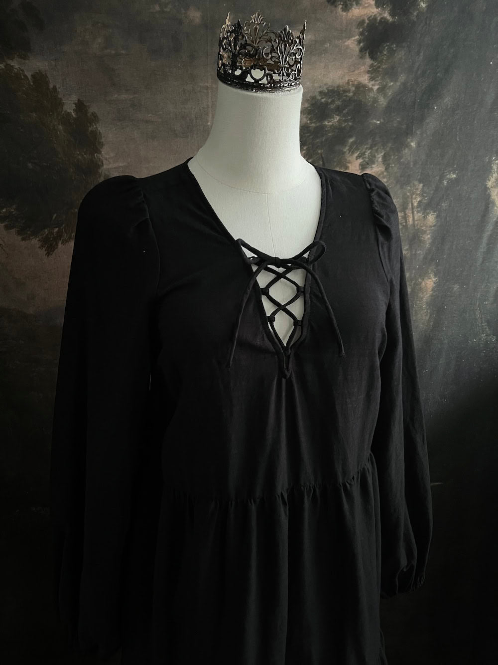 Historically inspired witchy gothic black tiered mini dress with corset lace up bodice and long bishop sleeves on a mannequin in front of a dark art history backdrop
