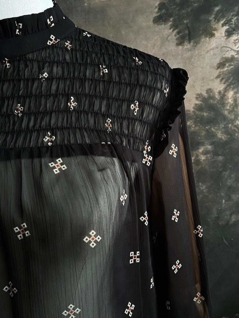 Mannequin wearing a historically-inspired yoked mock neck black blouse with cross patterns against a dark, forested painting background.