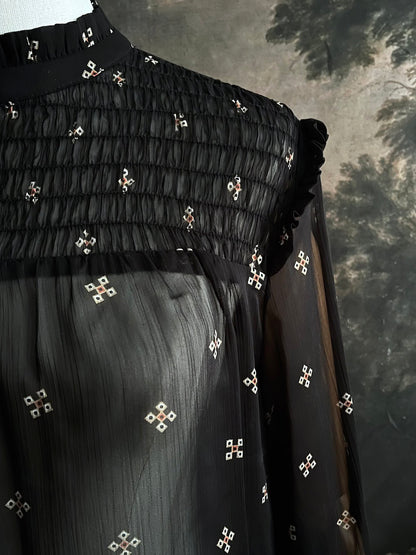 Mannequin wearing a historically-inspired yoked mock neck black blouse with cross patterns against a dark, forested painting background.