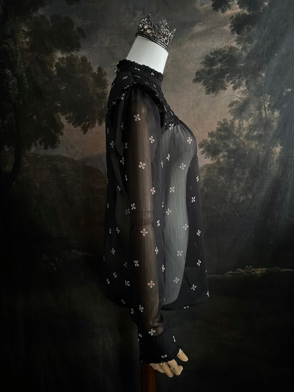 Mannequin wearing a historically-inspired yoked mock neck black blouse with cross patterns against a dark, forested painting background.