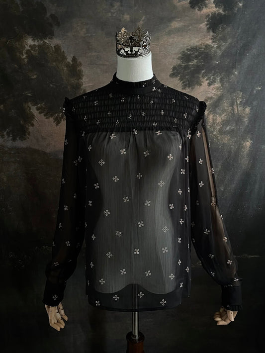 Mannequin wearing a historically-inspired yoked mock neck black blouse with cross patterns against a dark, forested painting background.