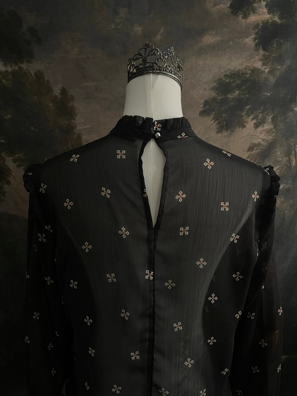 Mannequin wearing a historically-inspired yoked mock neck black blouse with cross patterns against a dark, forested painting background.