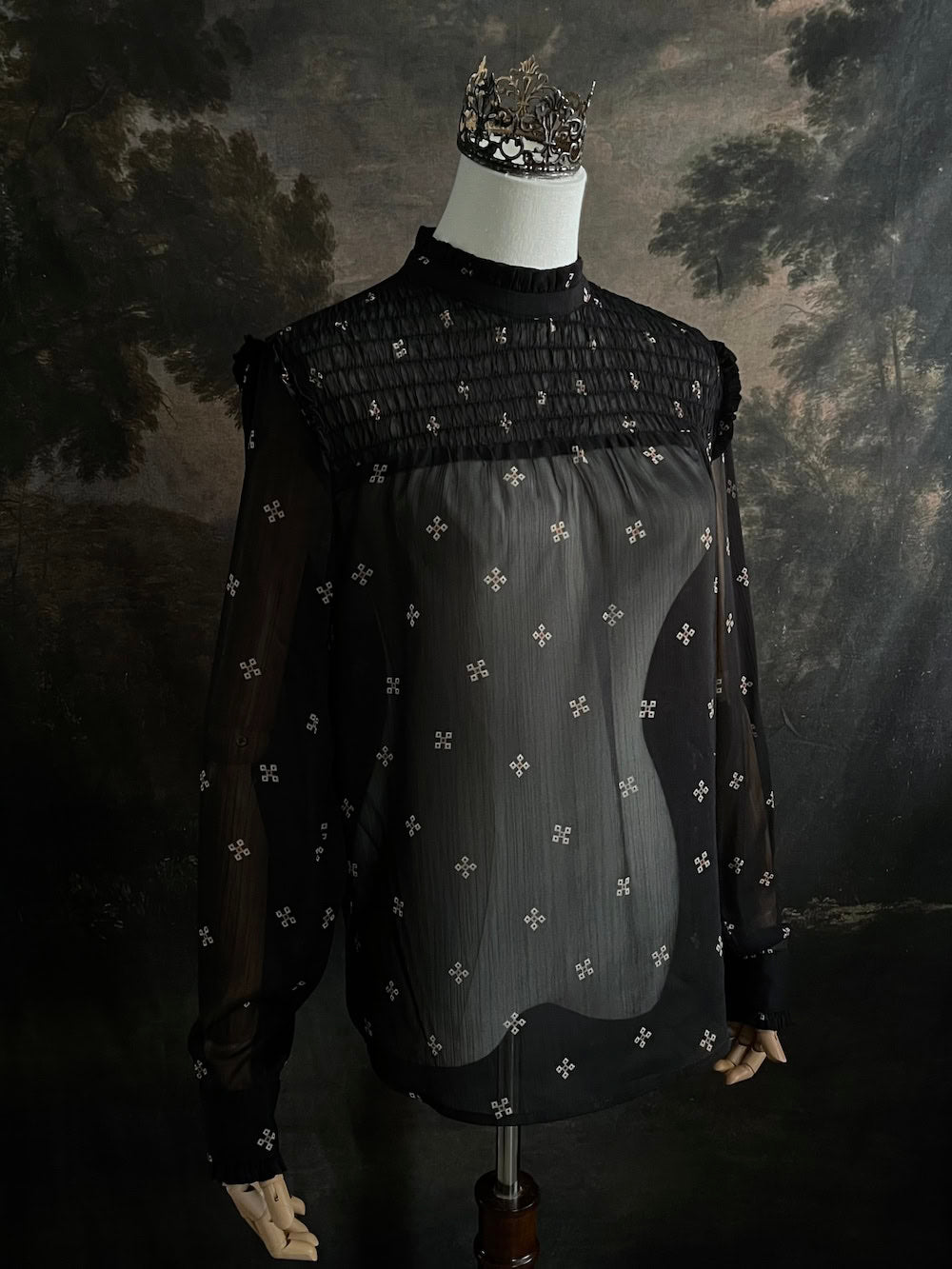 Mannequin wearing a historically-inspired yoked mock neck black blouse with cross patterns against a dark, forested painting background.