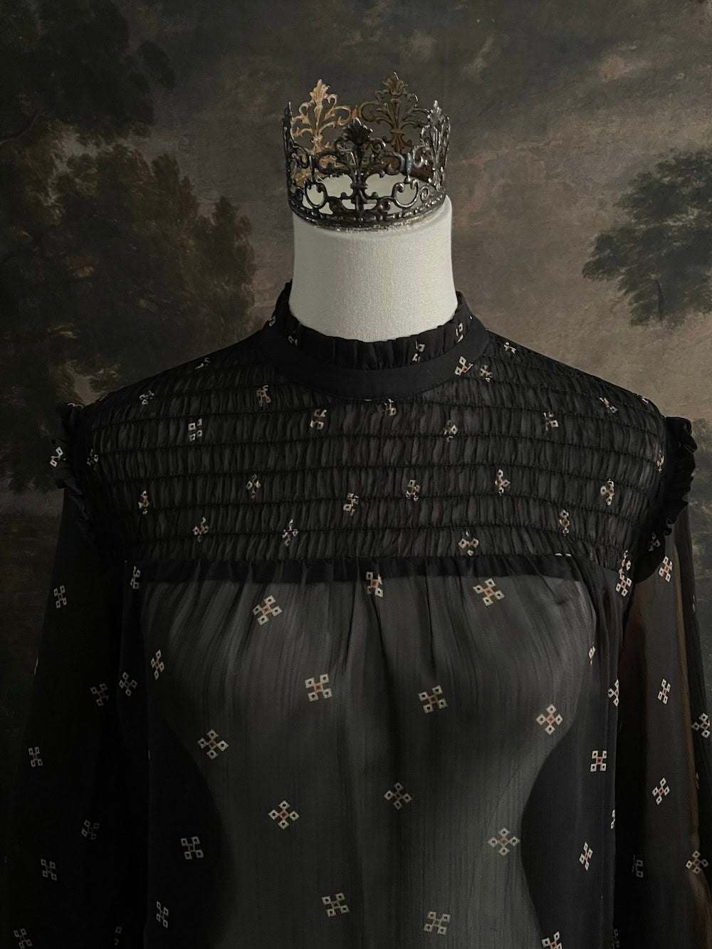 Mannequin wearing a historically-inspired yoked mock neck black blouse with cross patterns against a dark, forested painting background.
