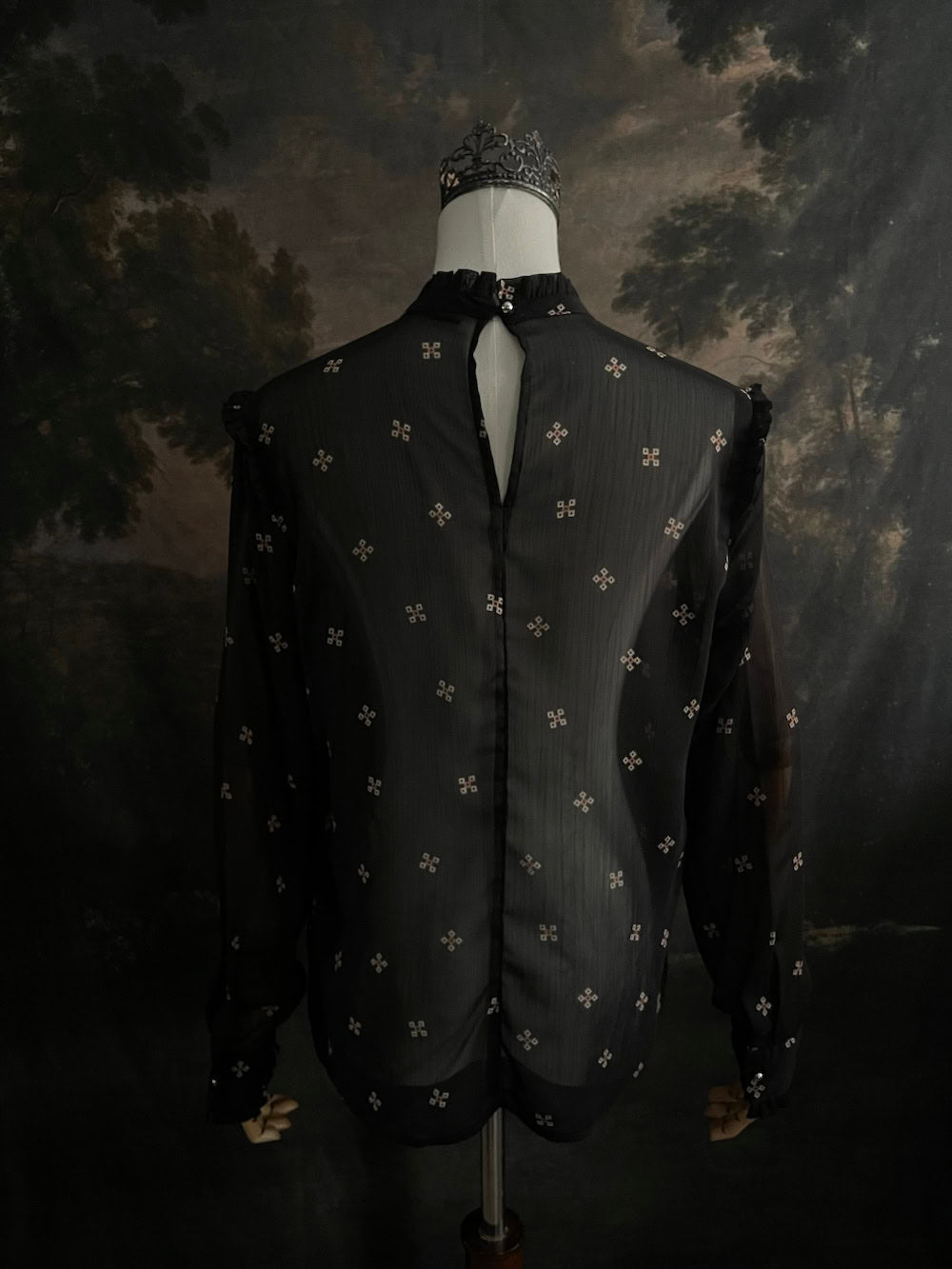 Mannequin wearing a historically-inspired yoked mock neck black blouse with cross patterns against a dark, forested painting background.