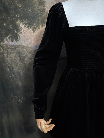 A Black Velvet Shirred Bodice Mini Dress inspired by the tudor and renaissance era on a mannequin in front of an art history backdrop