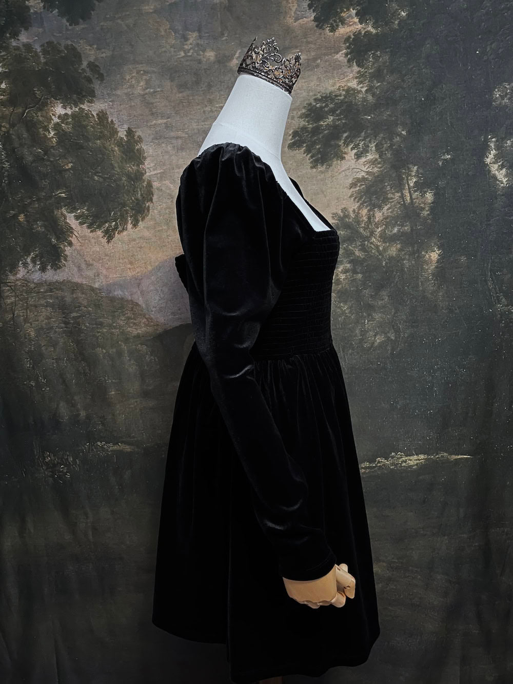 A Black Velvet Shirred Bodice Mini Dress inspired by the tudor and renaissance era on a mannequin in front of an art history backdrop