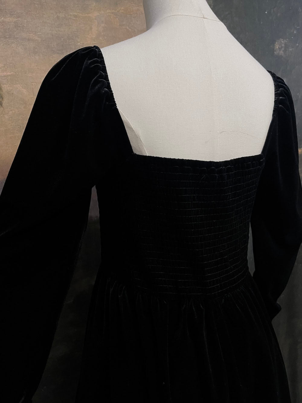 A Black Velvet Shirred Bodice Mini Dress inspired by the tudor and renaissance era on a mannequin in front of an art history backdrop