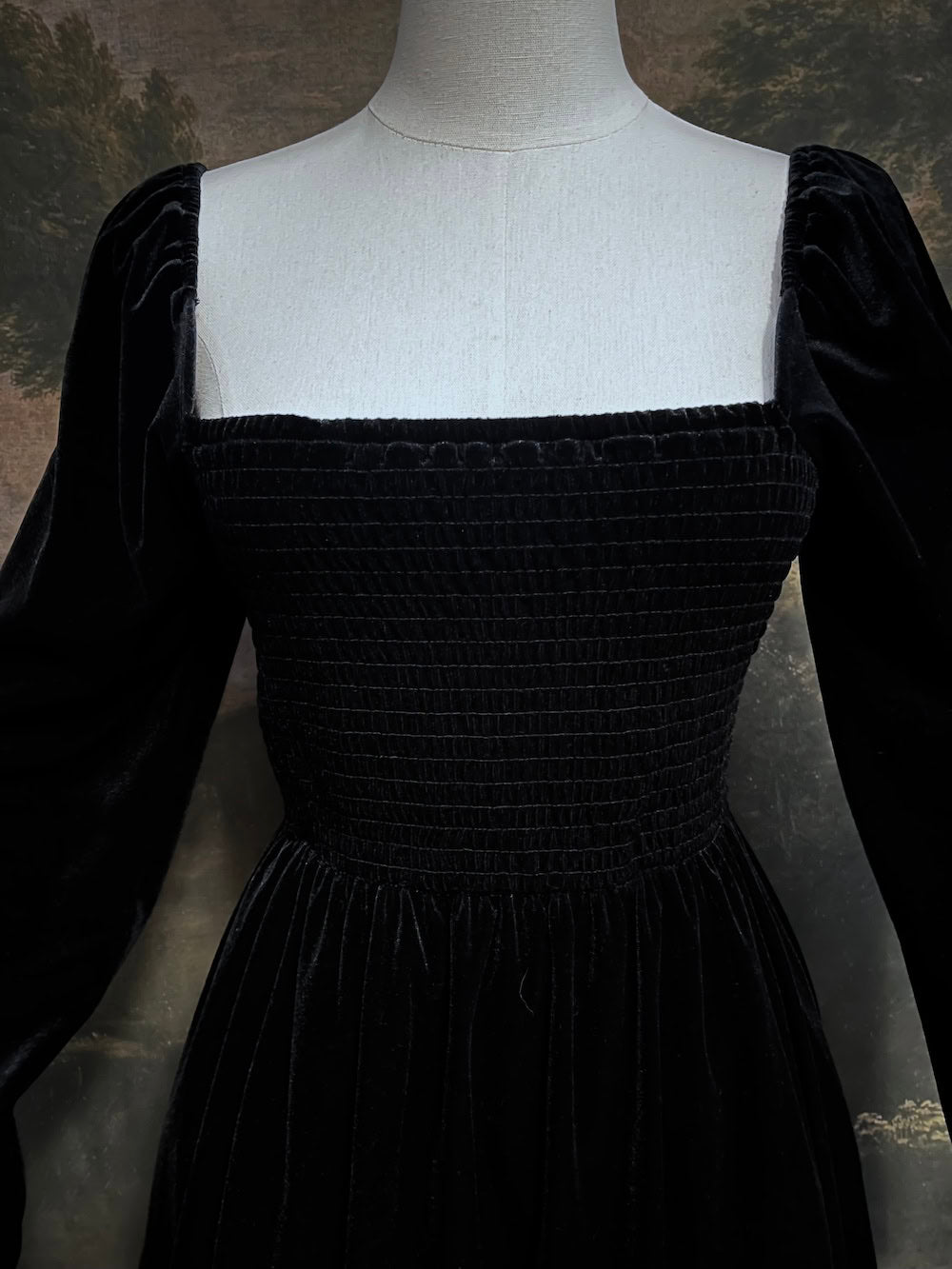 A Black Velvet Shirred Bodice Mini Dress inspired by the tudor and renaissance era on a mannequin in front of an art history backdrop