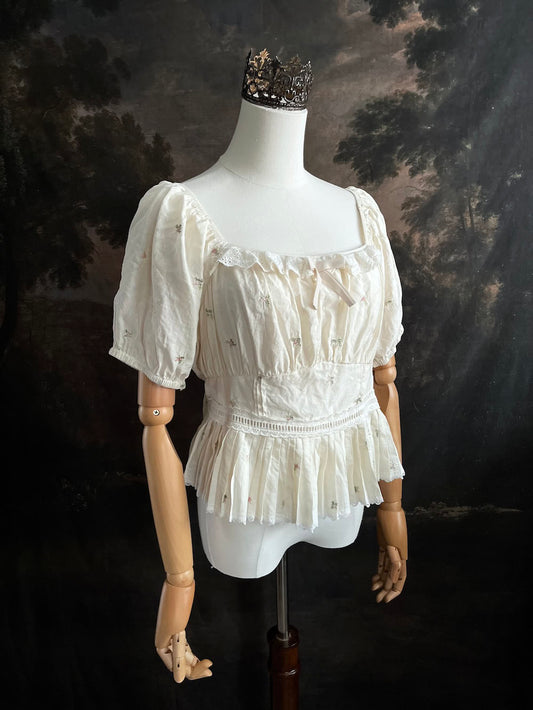 A vintage inspired historical fashion peplum top with embroidered floral designs and a pleated waistline on a mannequin in front of an art history backdrop
