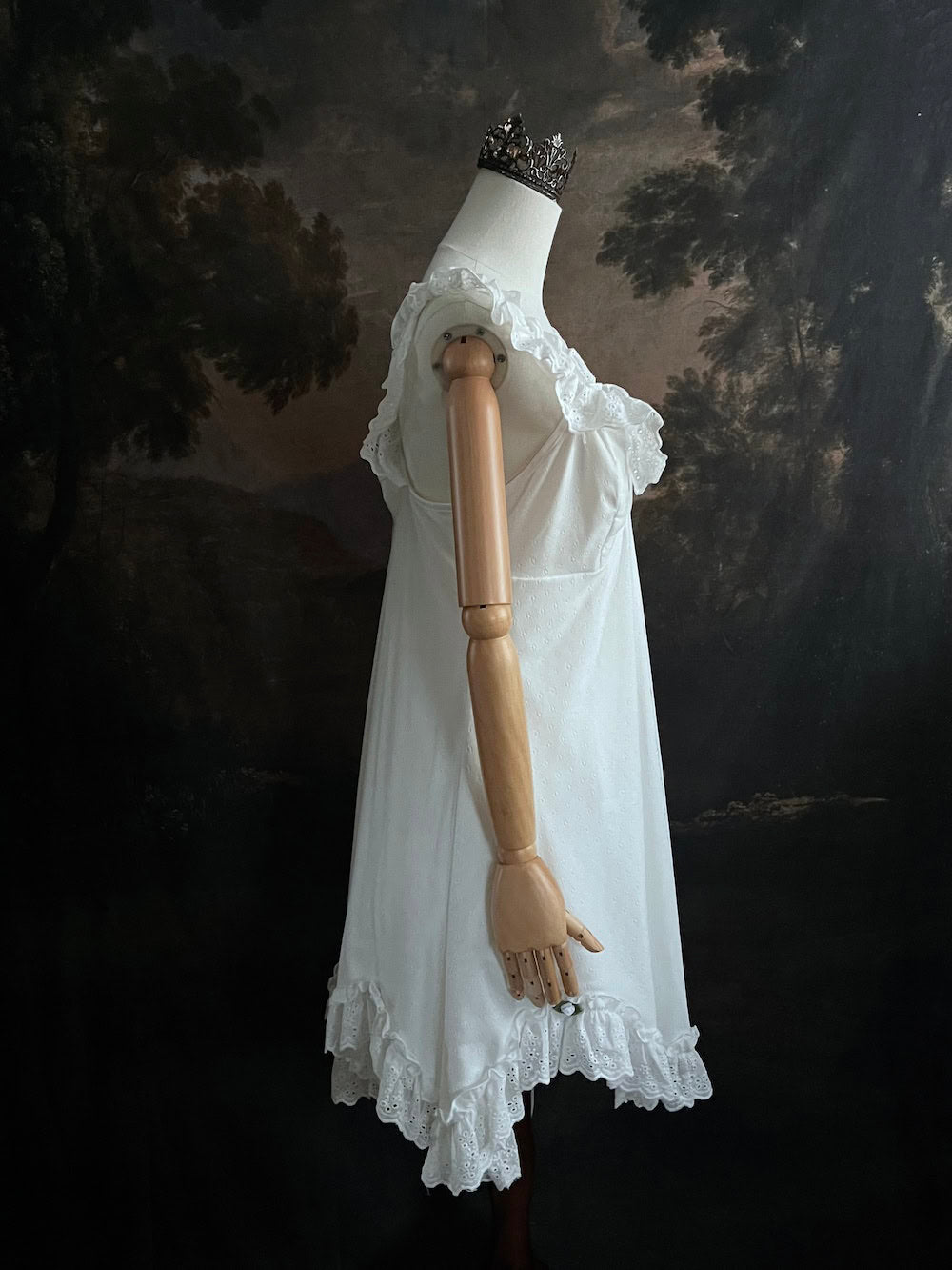 White Edwardian-inspired vintage style lace dress on a mannequin against a dark, forest-like art history background