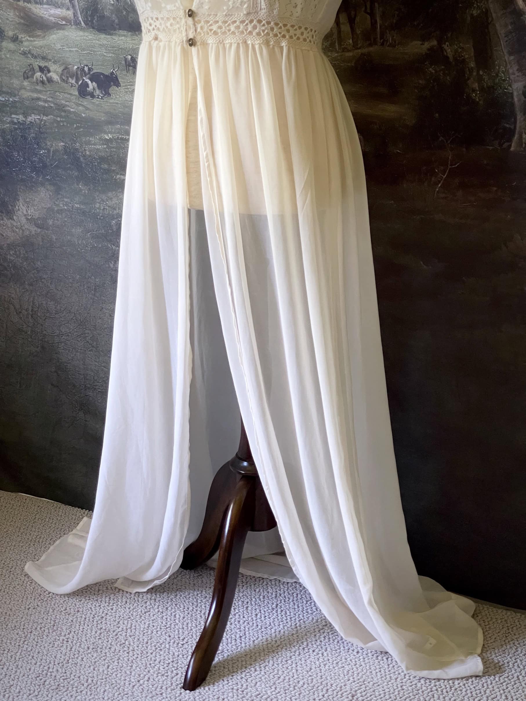 A historically inspired Edwardian era Ivory maxi Overdress with Floral Lace Details is pictured on a mannequin in front of an art history backdrop.