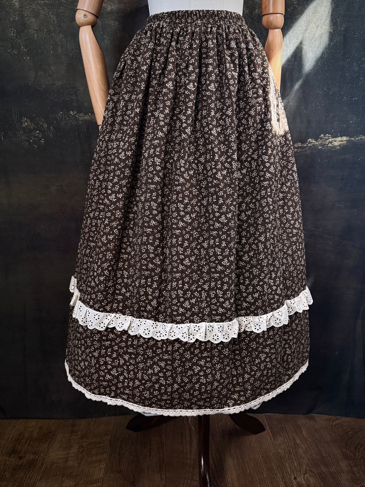 A victorian inspired dark brown floral print maxi skirt with white lace trim, displayed on a mannequin against a dark classical painting backdrop.