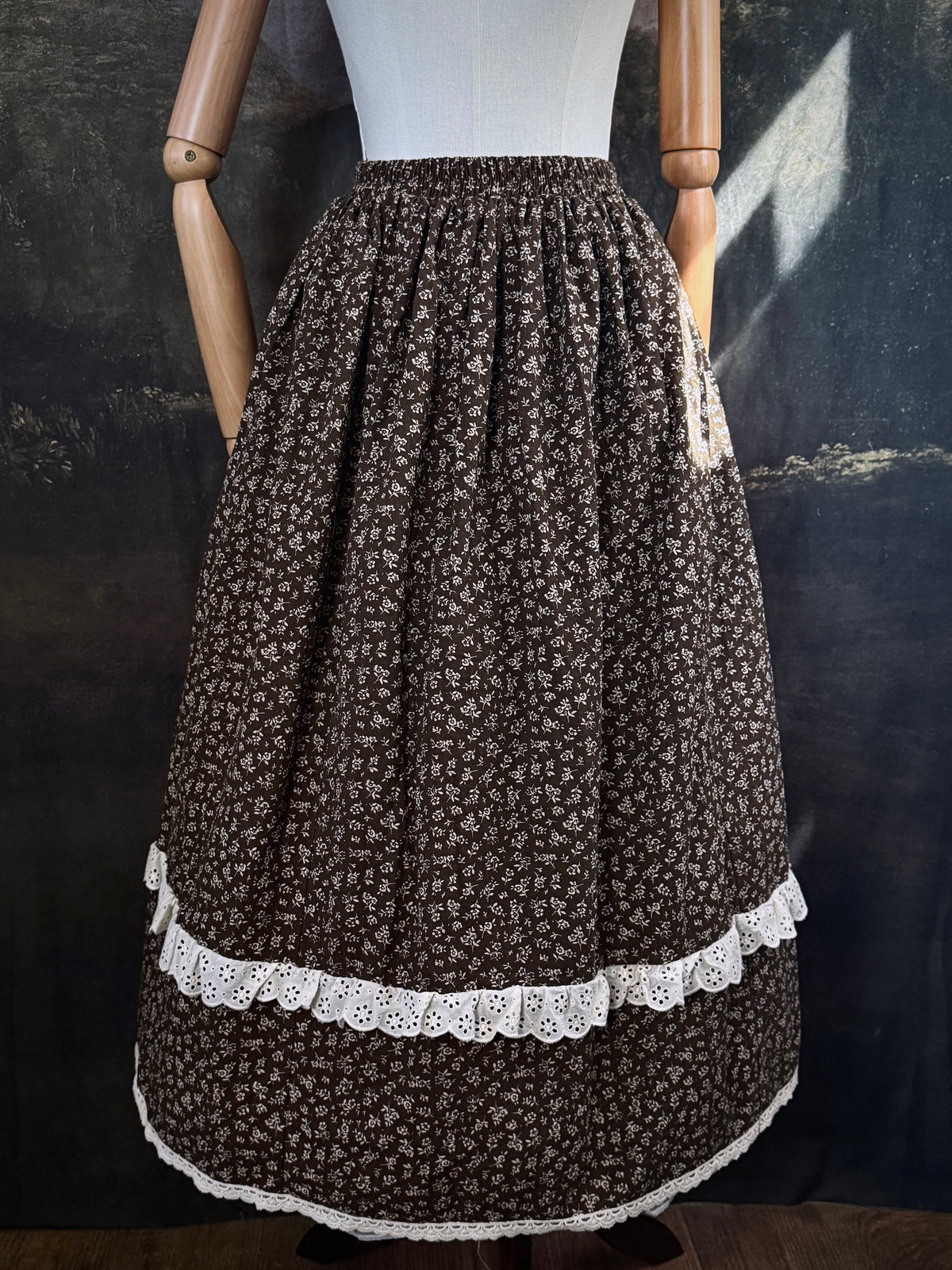 A victorian inspired dark brown floral print maxi skirt with white lace trim, displayed on a mannequin against a dark classical painting backdrop.
