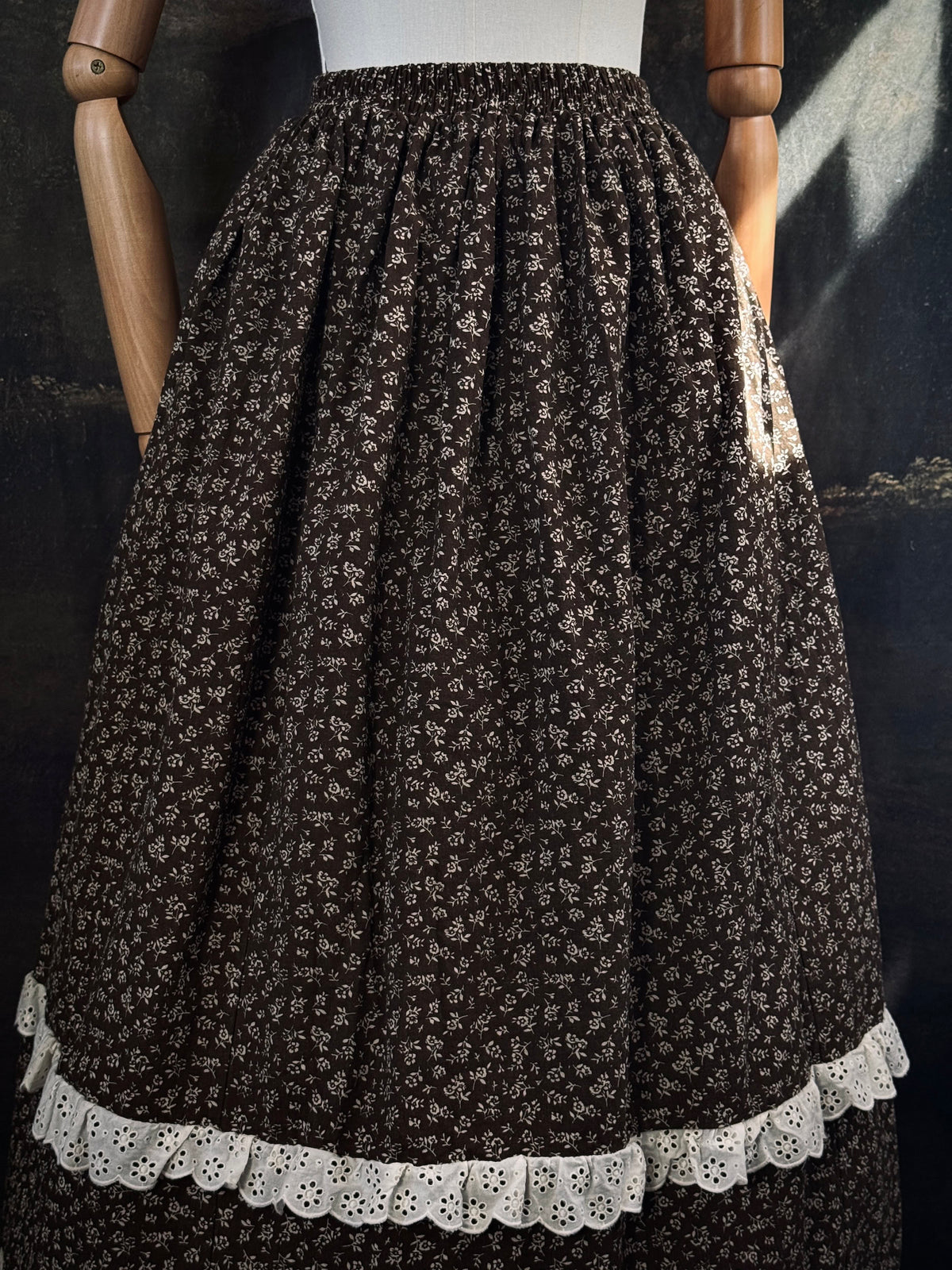 A victorian inspired dark brown floral print maxi skirt with white lace trim, displayed on a mannequin against a dark classical painting backdrop.