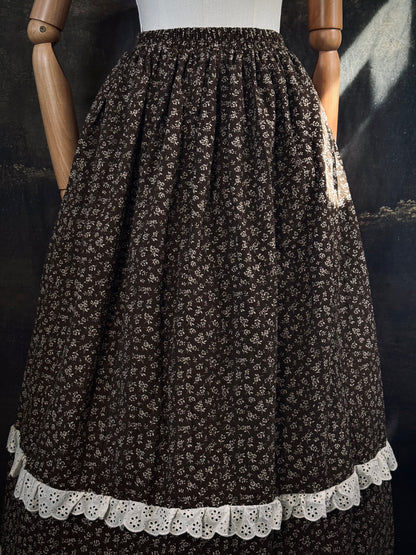 A victorian inspired dark brown floral print maxi skirt with white lace trim, displayed on a mannequin against a dark classical painting backdrop.