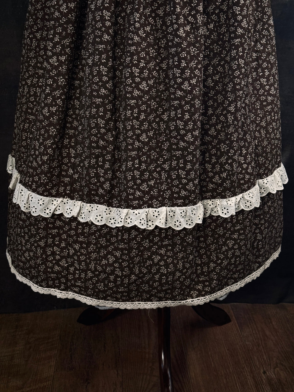 A victorian inspired dark brown floral print maxi skirt with white lace trim, displayed on a mannequin against a dark classical painting backdrop.