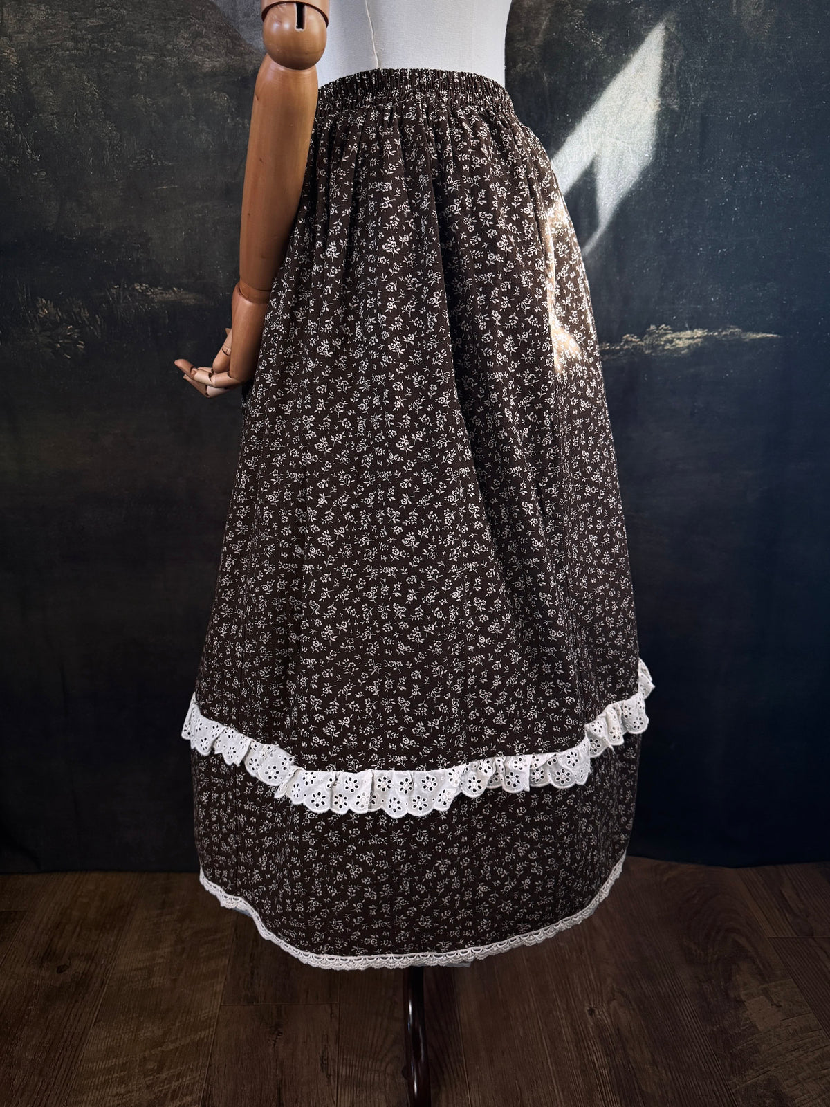 A victorian inspired dark brown floral print maxi skirt with white lace trim, displayed on a mannequin against a dark classical painting backdrop.