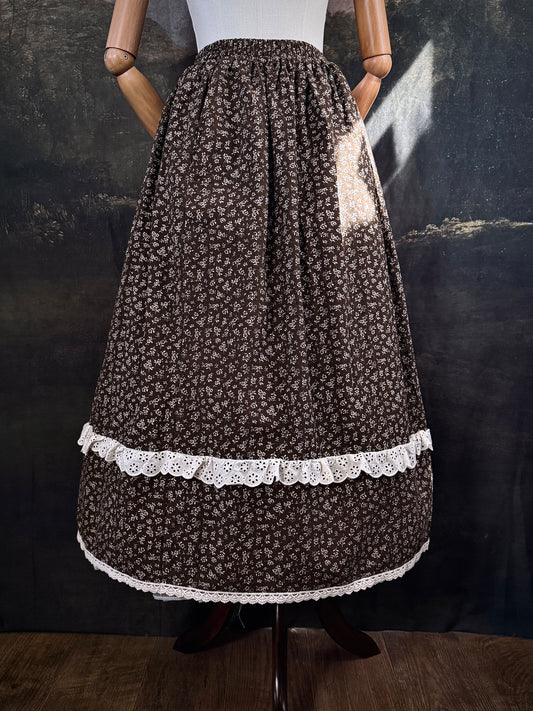 A victorian inspired dark brown floral print maxi skirt with white lace trim, displayed on a mannequin against a dark classical painting backdrop.