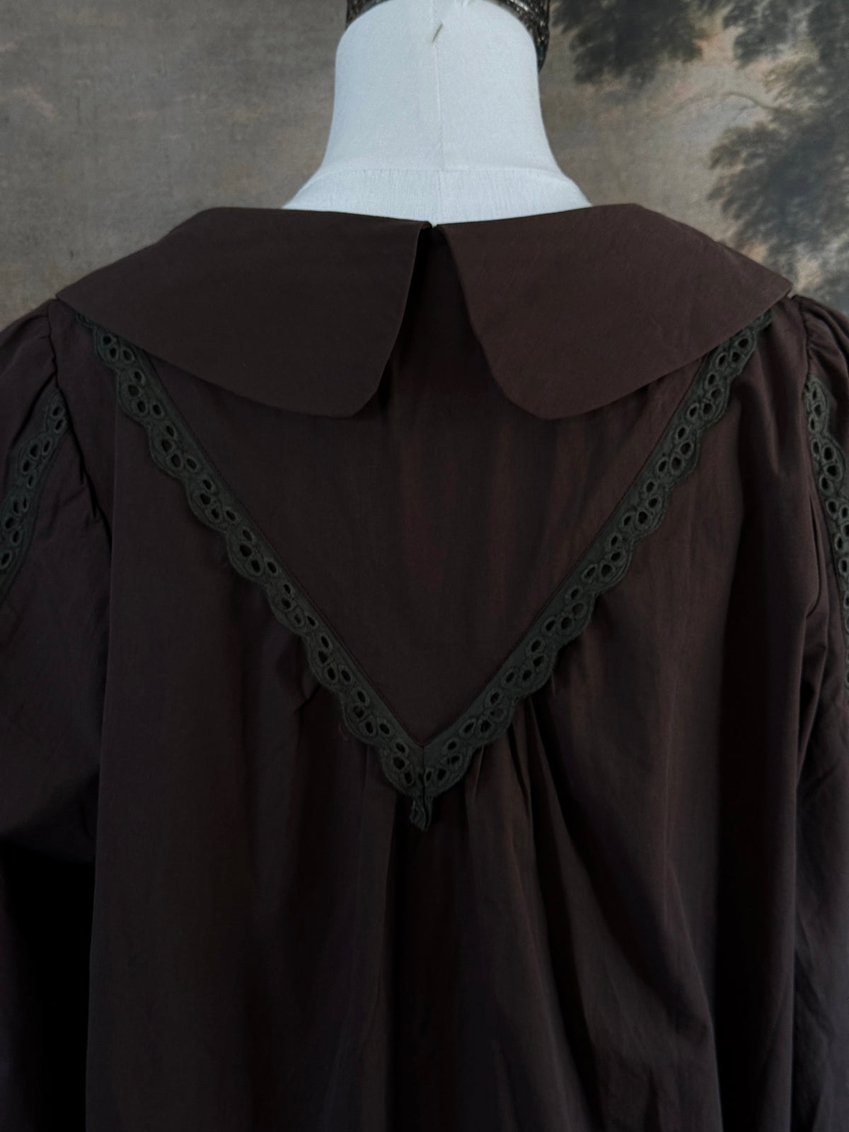 A historically inspired victorian era dark brown long-sleeve blouse with a large Peter Pan collar and lace trim details, displayed on a mannequin against a dark forest landscape backdrop.