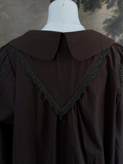 A historically inspired victorian era dark brown long-sleeve blouse with a large Peter Pan collar and lace trim details, displayed on a mannequin against a dark forest landscape backdrop.