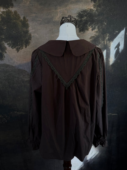 A historically inspired victorian era dark brown long-sleeve blouse with a large Peter Pan collar and lace trim details, displayed on a mannequin against a dark forest landscape backdrop.