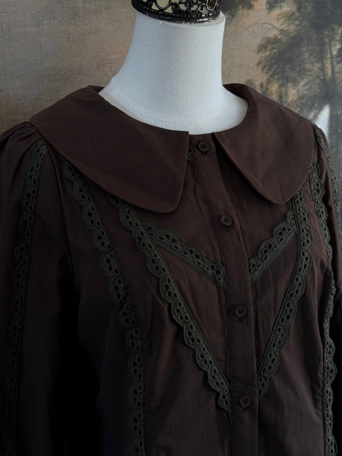 A historically inspired victorian era dark brown long-sleeve blouse with a large Peter Pan collar and lace trim details, displayed on a mannequin against a dark forest landscape backdrop.