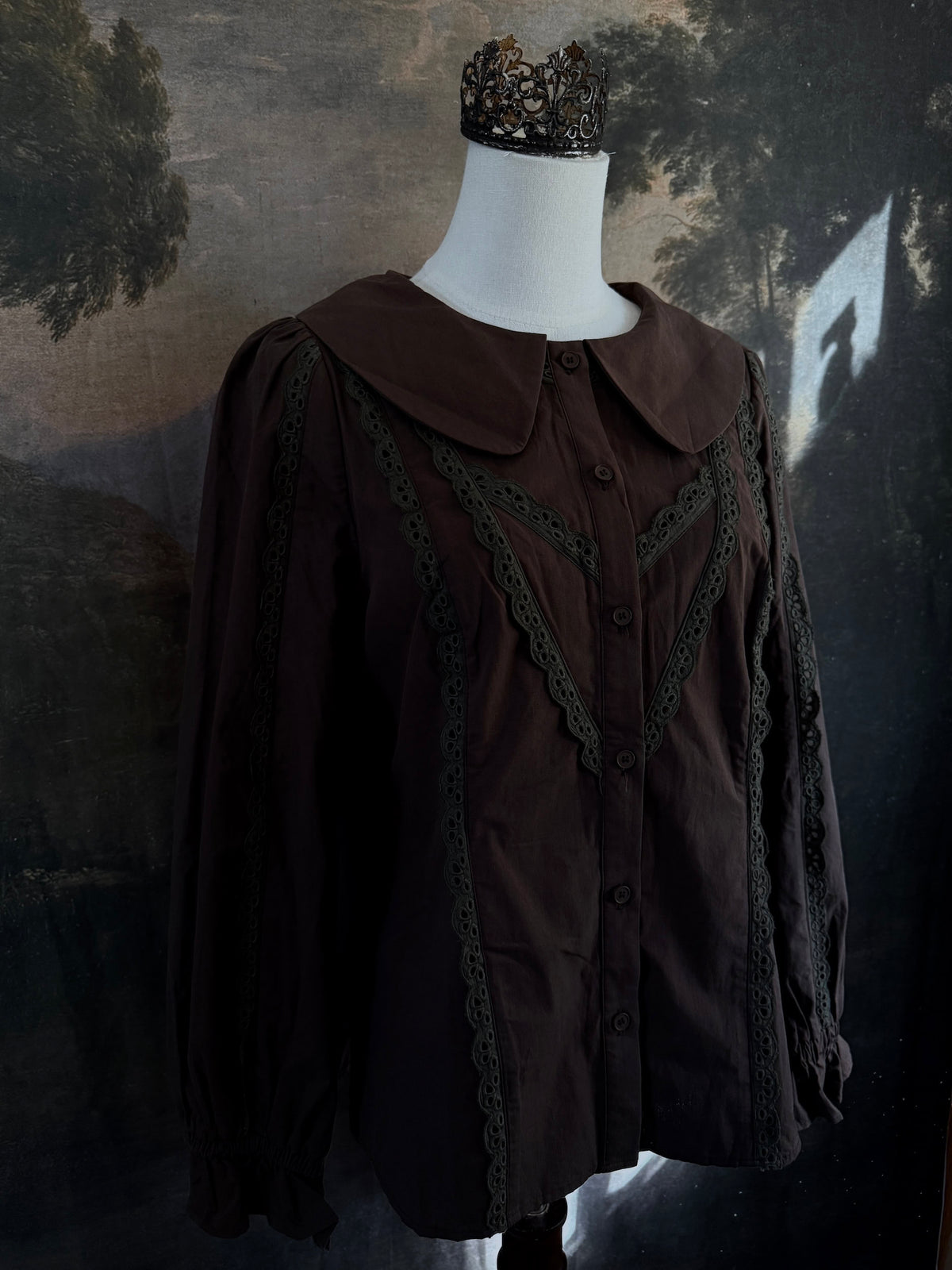 A historically inspired victorian era dark brown long-sleeve blouse with a large Peter Pan collar and lace trim details, displayed on a mannequin against a dark forest landscape backdrop.