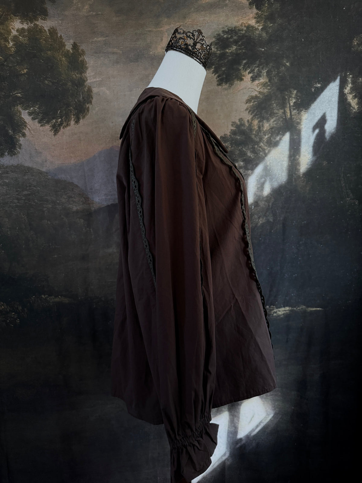 A historically inspired victorian era dark brown long-sleeve blouse with a large Peter Pan collar and lace trim details, displayed on a mannequin against a dark forest landscape backdrop.