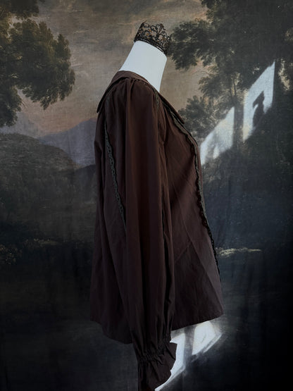 A historically inspired victorian era dark brown long-sleeve blouse with a large Peter Pan collar and lace trim details, displayed on a mannequin against a dark forest landscape backdrop.