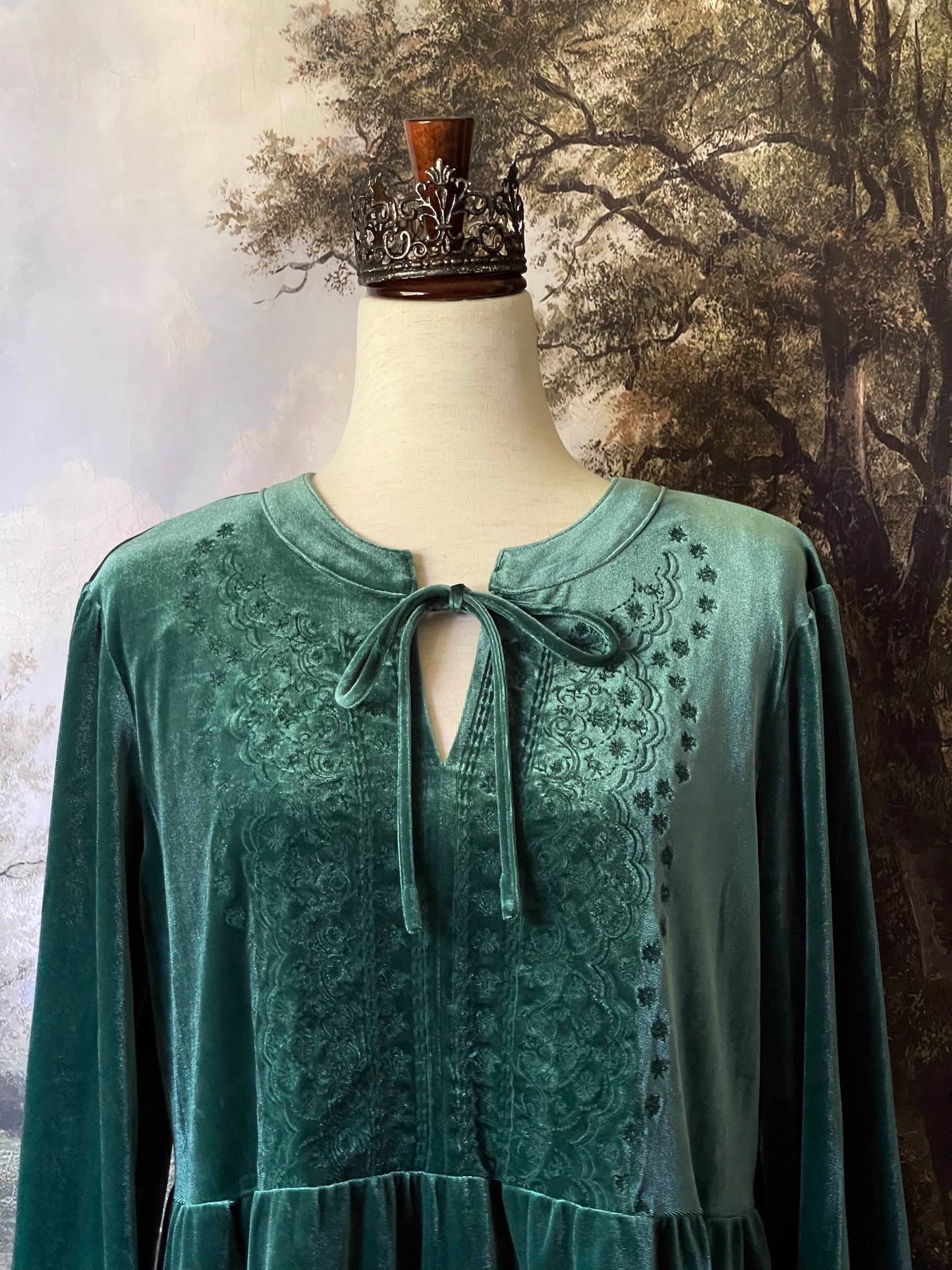 A Medieval Fantasy Floral Embroidery Velour Tunic in Emerald Green is pictured on a mannequin in front of a forested fantasy backdrop.