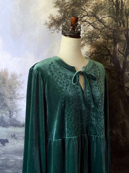 A Medieval Fantasy Floral Embroidery Velour Tunic in Emerald Green is pictured on a mannequin in front of a forested fantasy backdrop.