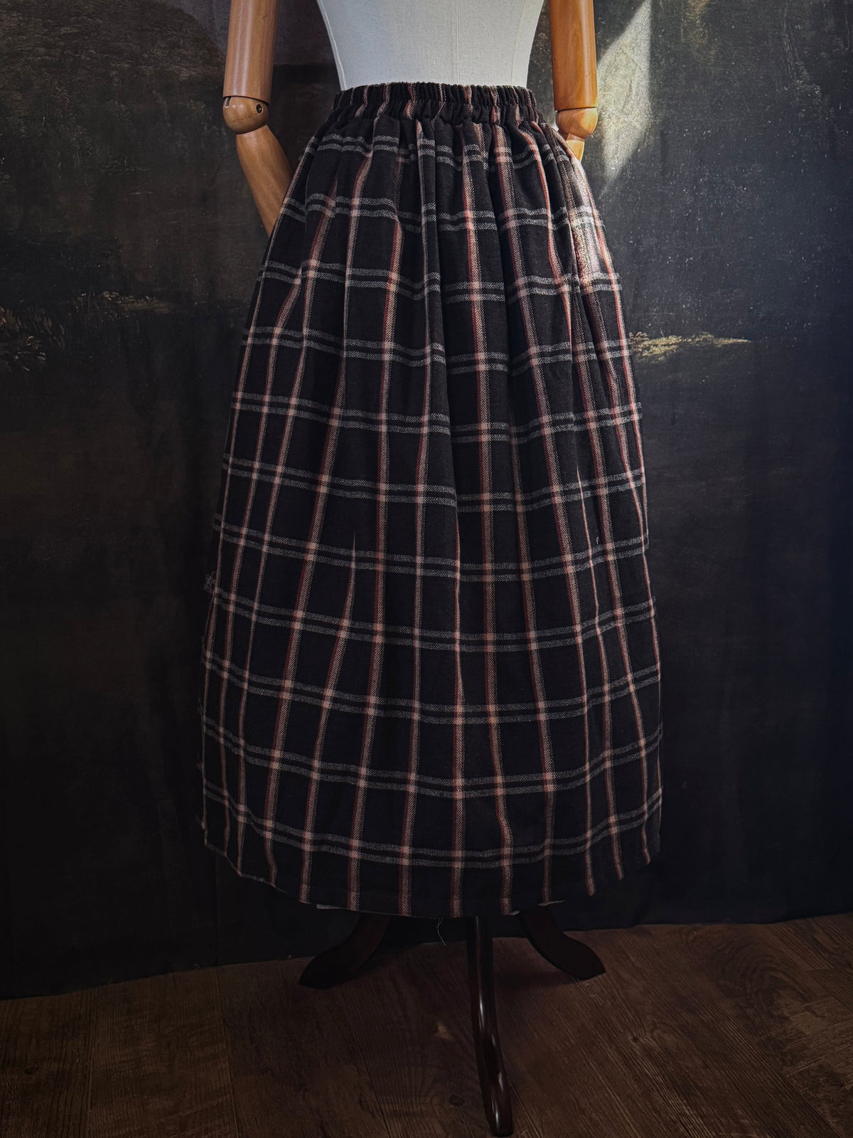 A dark brown and beige and rust colored autumnal herringbone woven tartan plaid skirt inspired by historical scotland, pictured on a mannequin in front of a dark forest landscape painting backdrop.