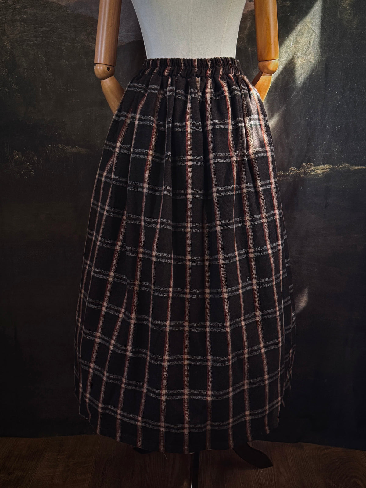 A dark brown and beige and rust colored autumnal herringbone woven tartan plaid skirt inspired by historical scotland, pictured on a mannequin in front of a dark forest landscape painting backdrop.