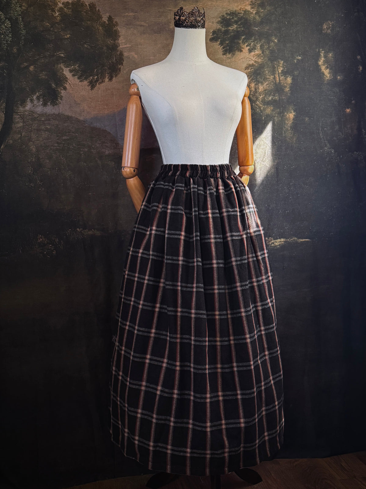 A dark brown and beige and rust colored autumnal herringbone woven tartan plaid skirt inspired by historical scotland, pictured on a mannequin in front of a dark forest landscape painting backdrop.