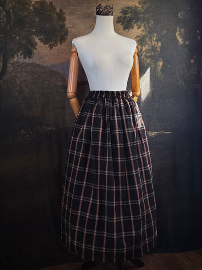 A dark brown and beige and rust colored autumnal herringbone woven tartan plaid skirt inspired by historical scotland, pictured on a mannequin in front of a dark forest landscape painting backdrop.