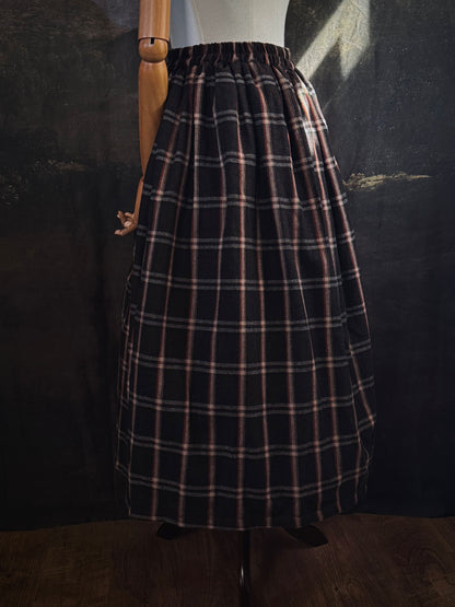 A dark brown and beige and rust colored autumnal herringbone woven tartan plaid skirt inspired by historical scotland, pictured on a mannequin in front of a dark forest landscape painting backdrop.