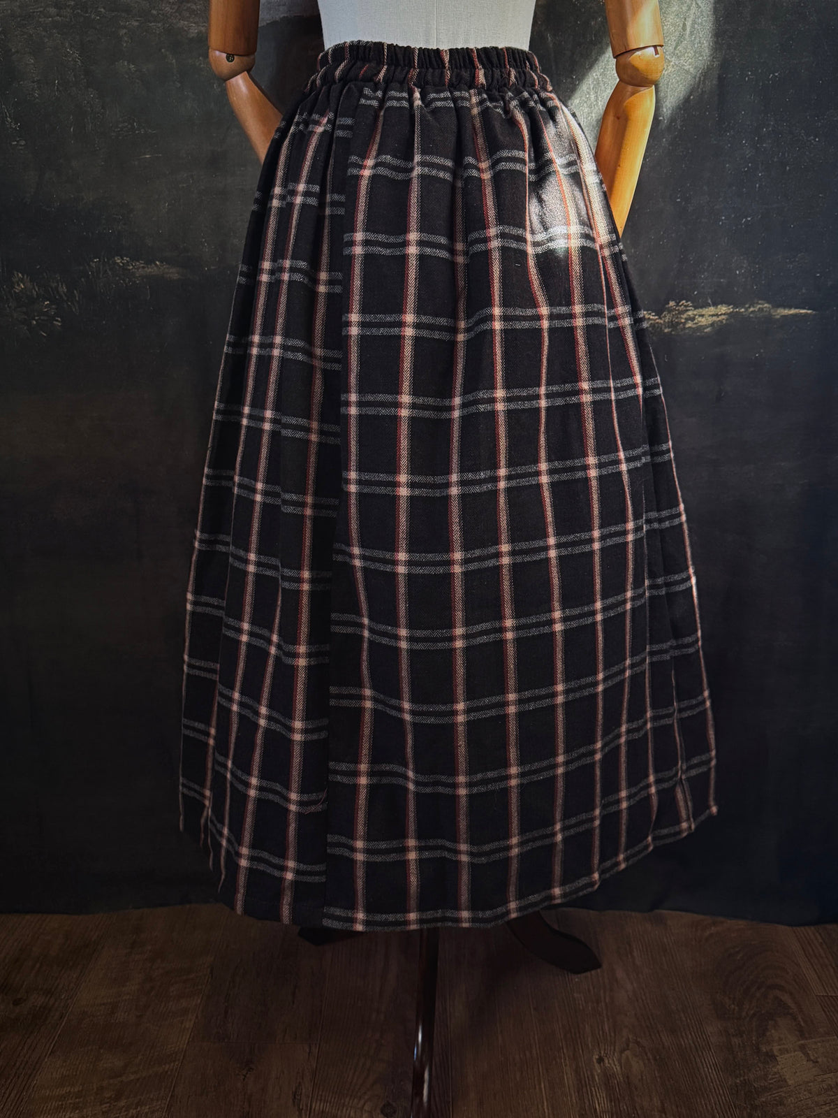 A dark brown and beige and rust colored autumnal herringbone woven tartan plaid skirt inspired by historical scotland, pictured on a mannequin in front of a dark forest landscape painting backdrop.