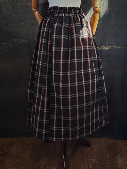 A dark brown and beige and rust colored autumnal herringbone woven tartan plaid skirt inspired by historical scotland, pictured on a mannequin in front of a dark forest landscape painting backdrop.